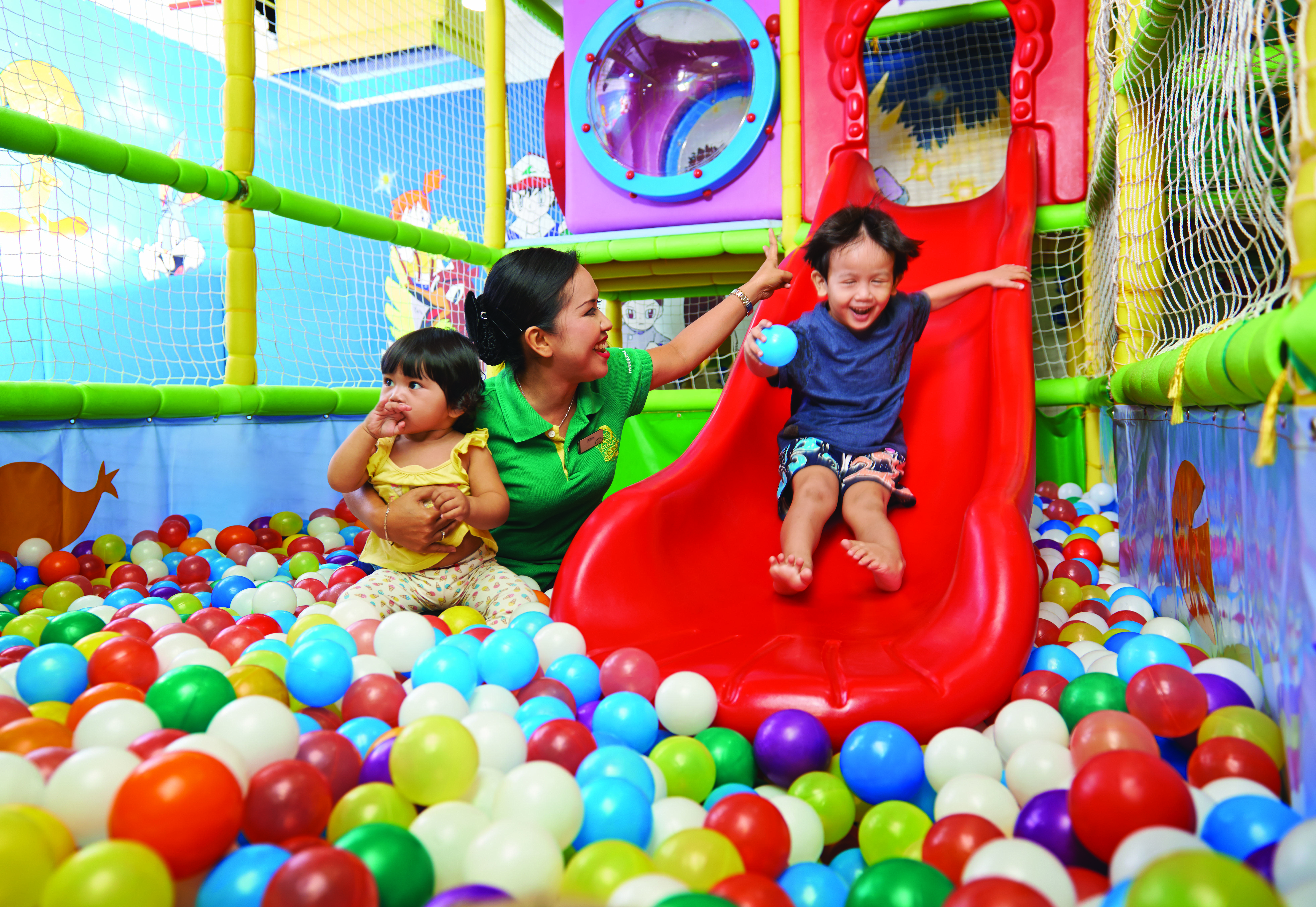 槟城宾乐雅度假村酒店 峇都丁宜 外观 照片 The photo shows a playful indoor environment designed for children. In the center, there is a red slide that a young boy is joyfully sliding down. He appears to be having fun, smiling as he descends. Nearby, a woman is attentively engaging with him, 