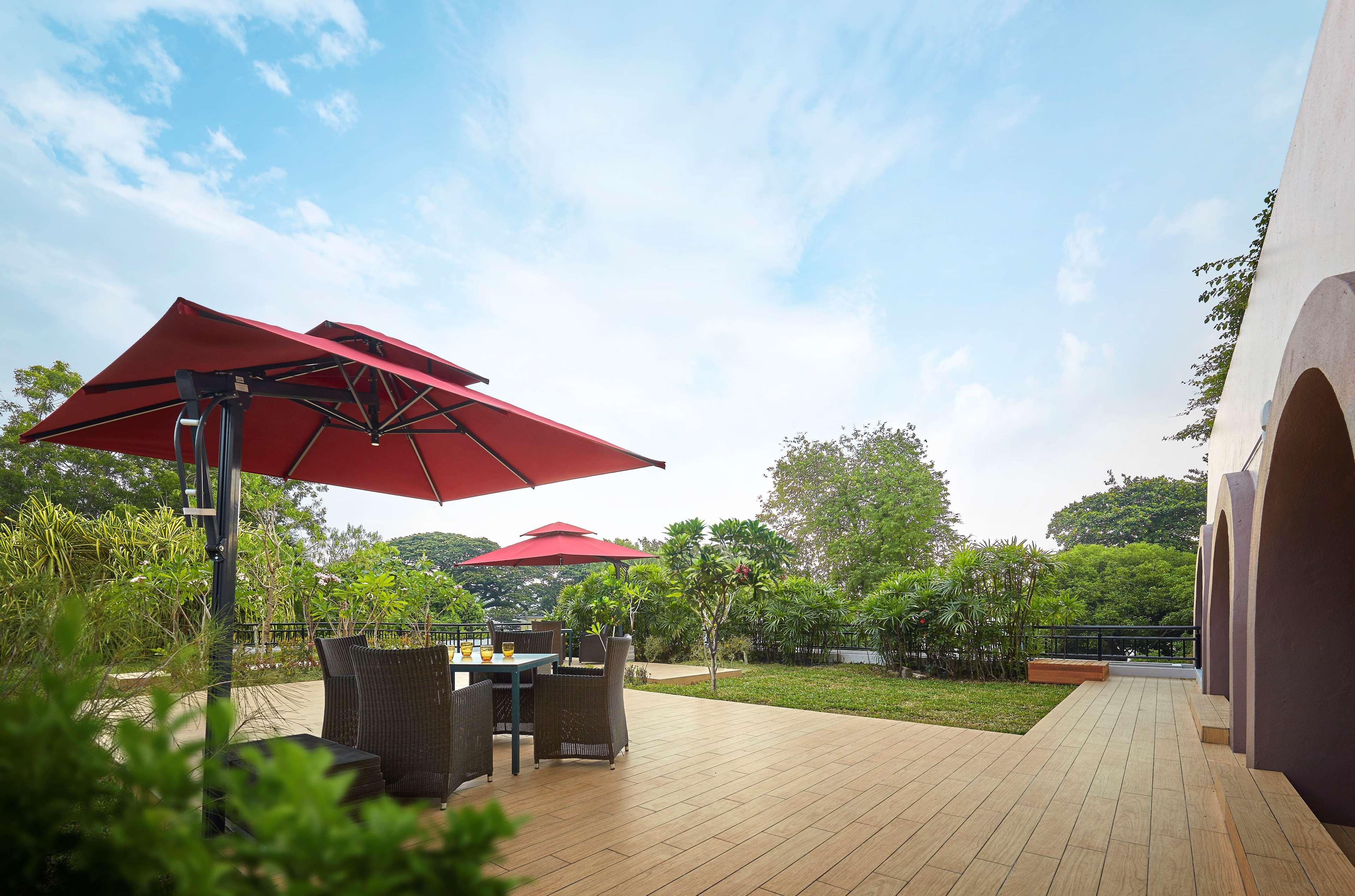 槟城宾乐雅度假村酒店 峇都丁宜 外观 照片 The photo shows an outdoor patio area with a wooden deck. There are two red umbrellas providing shade over a small dining table with dark chairs. Surrounding the area are lush green plants and trees, contributing to a serene atmosphere. The sky above