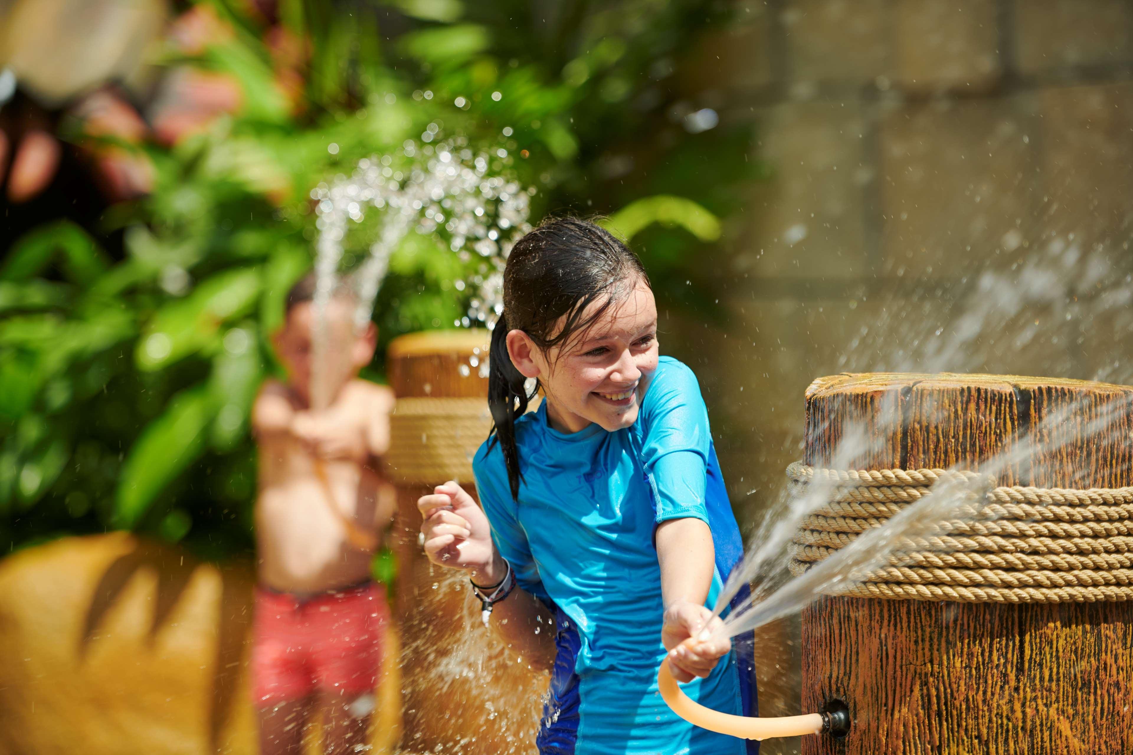 槟城宾乐雅度假村酒店 峇都丁宜 外观 照片 The photo depicts a joyful scene at a water park or a similar environment. In the foreground, a girl wearing a blue swimsuit is smiling as she interacts with a water feature that sprays water. She appears to be having a lot of fun. In the background,