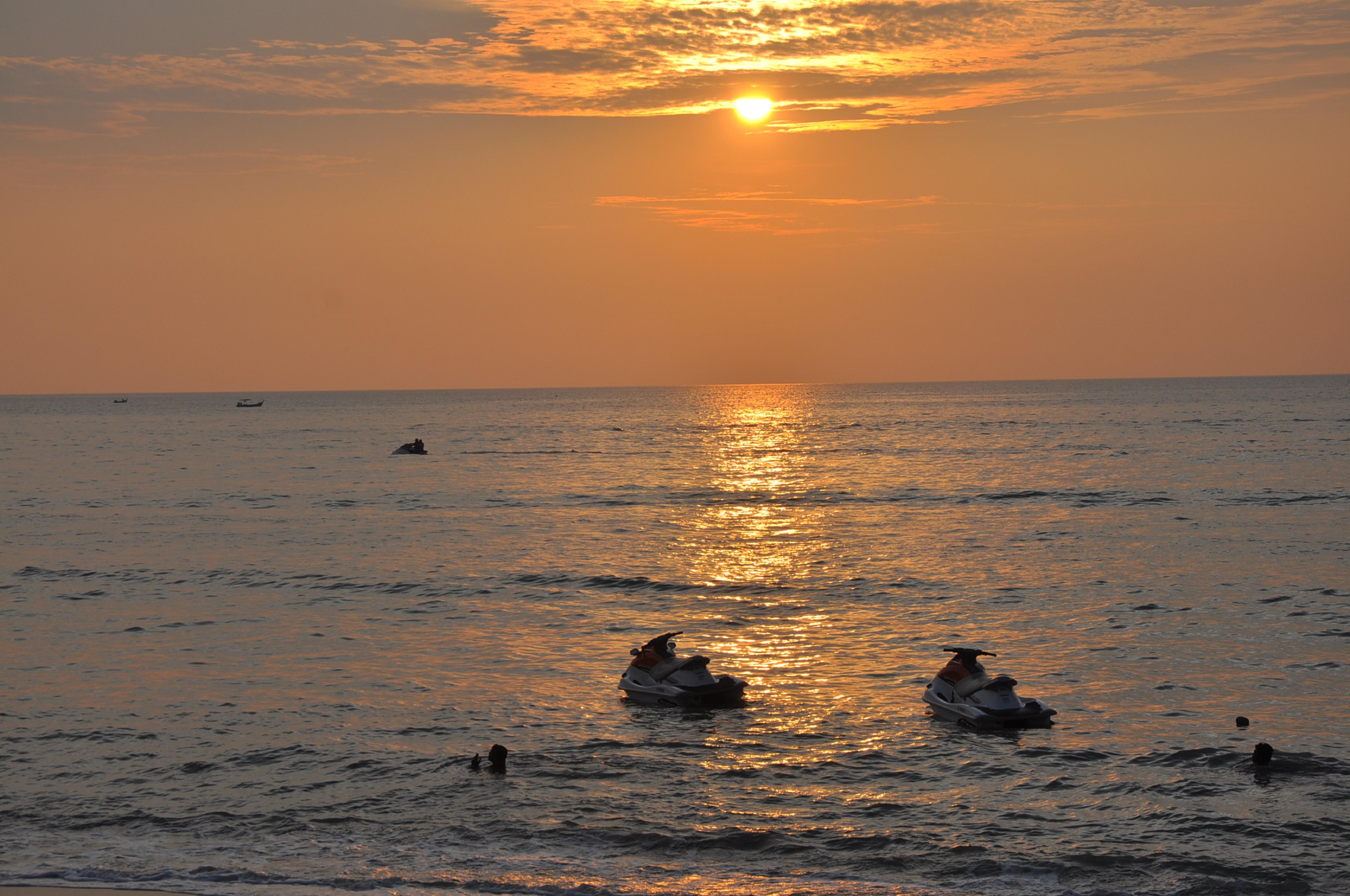 槟城宾乐雅度假村酒店 峇都丁宜 外观 照片 The photo depicts a serene sunset over the ocean. The sun is low on the horizon, casting golden and orange hues across the sky and reflecting on the water's surface. There are a couple of jetskis visible in the foreground, with individuals riding the