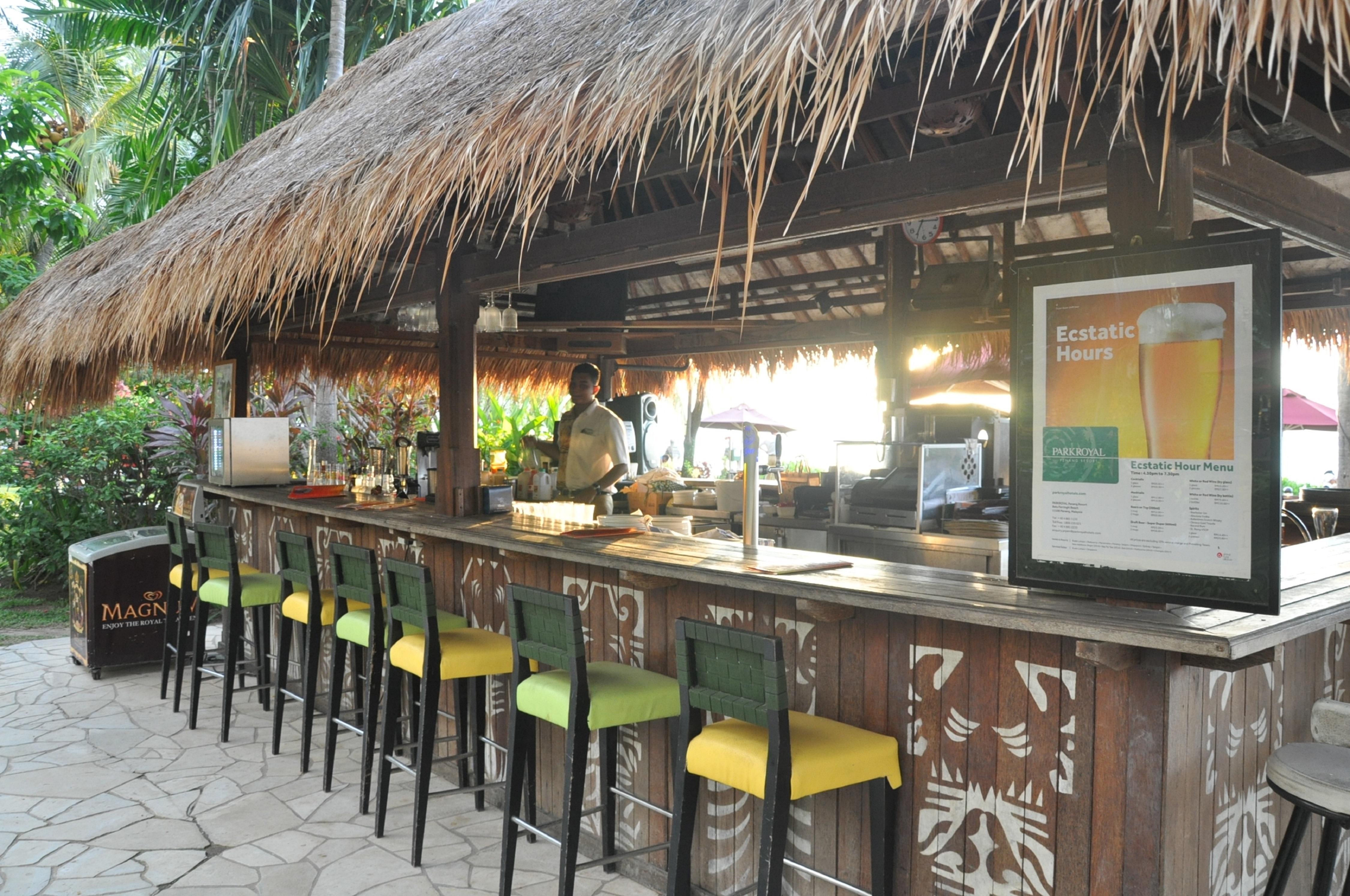 槟城宾乐雅度假村酒店 峇都丁宜 外观 照片 The photo shows an outdoor bar with a thatched roof, characteristic of a tropical or resort setting. The bar is made of wood and features several bar stools with green and yellow seats. Behind the bar, a bartender is preparing drinks, with various in